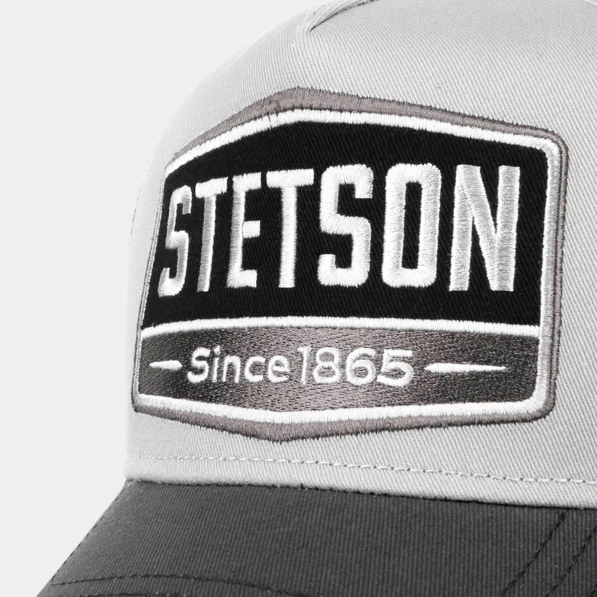 Gros plan sur l'écusson Stetson Since 1865 brodé noir et gris de la casquette trucker Highway, devant gris clair