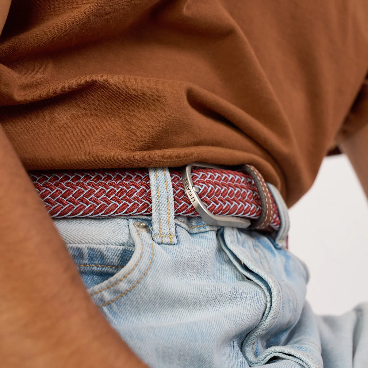 Boucle argentée gravée BillyBelt refermée sur le tressage rouge brique et bleu ciel de la ceinture Sienne, sur un jean clair