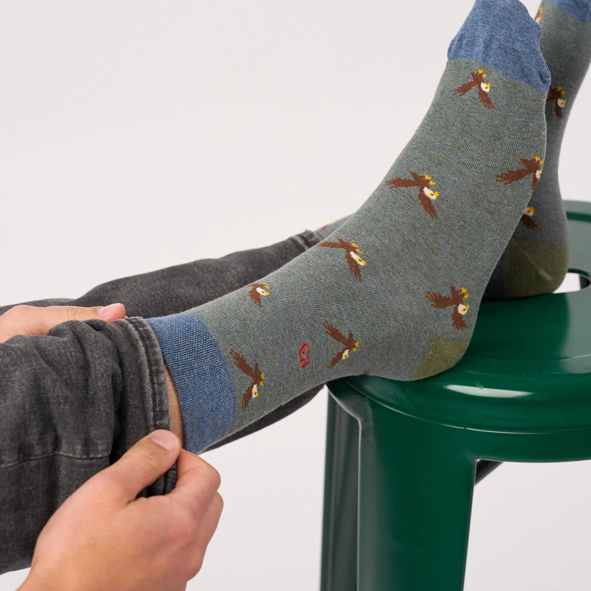 Chaussettes Aigle BillyBelt portées pieds posés sur un tabouret vert, motif aigles et logo poisson brodé rouge