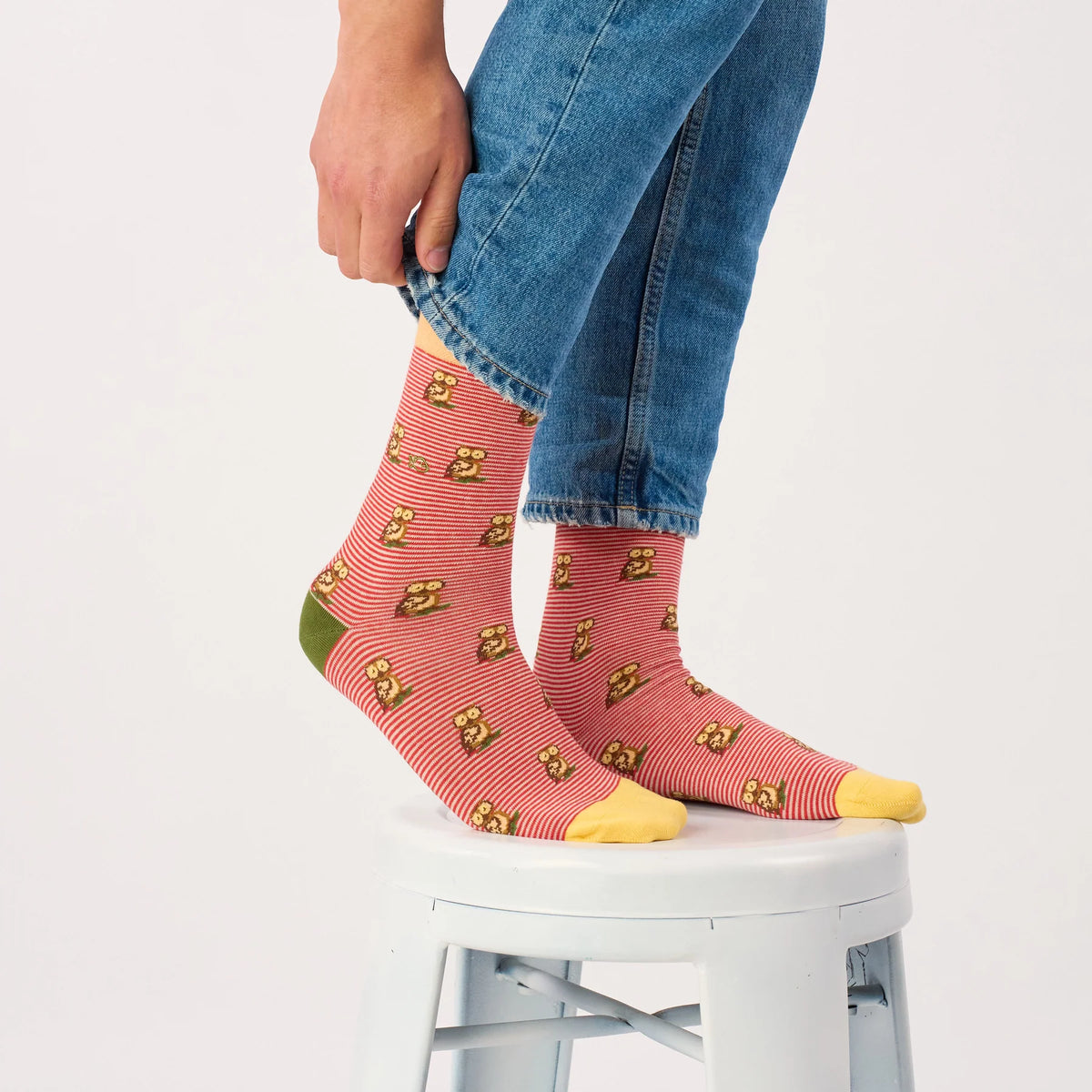 Paire de chaussettes Hibou rouges portées sur un tabouret blanc, jean retroussé à la main au-dessus de la tige mi-mollet