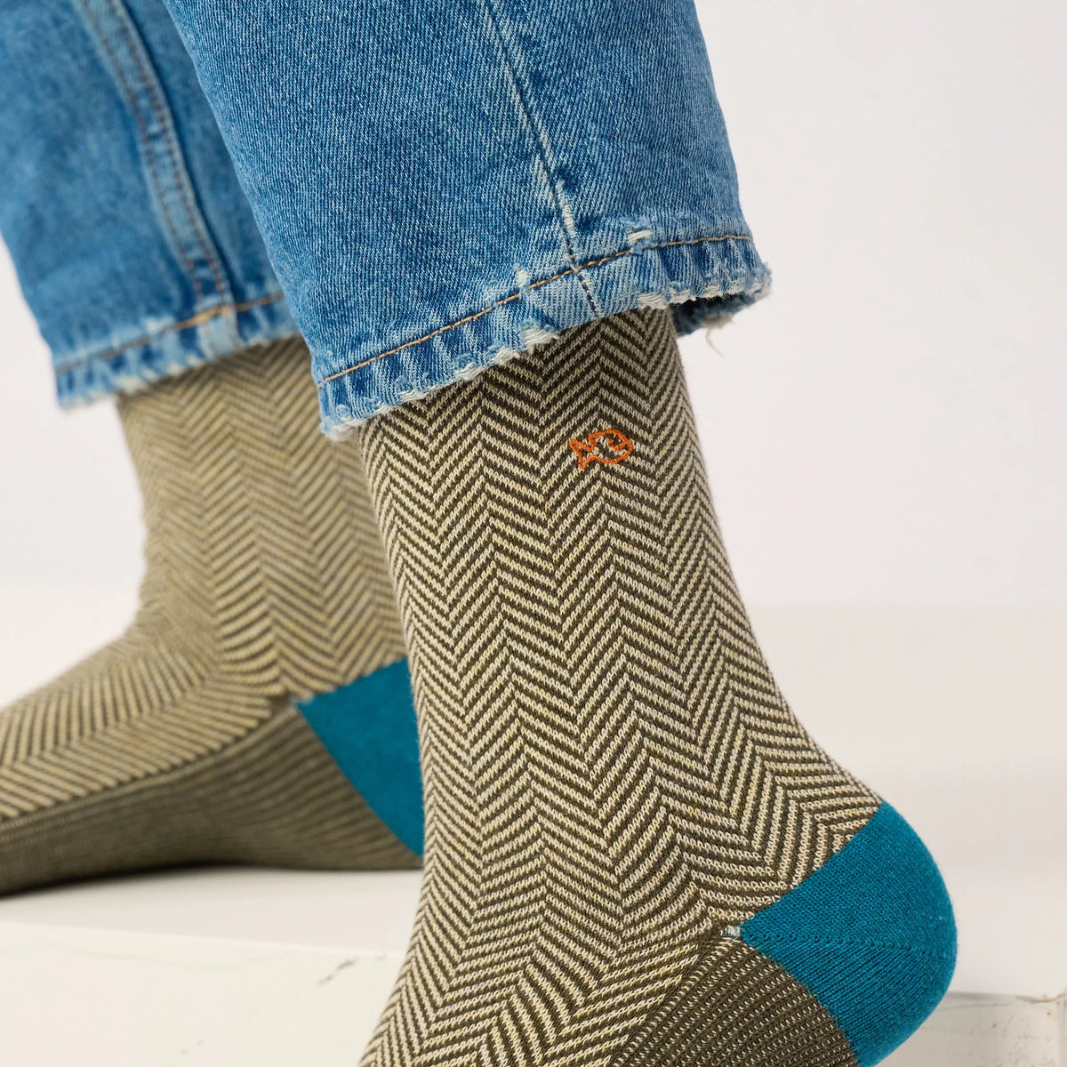 Gros plan sur le chevron kaki et écru sous un ourlet de jean, talon bleu canard et logo poisson orange