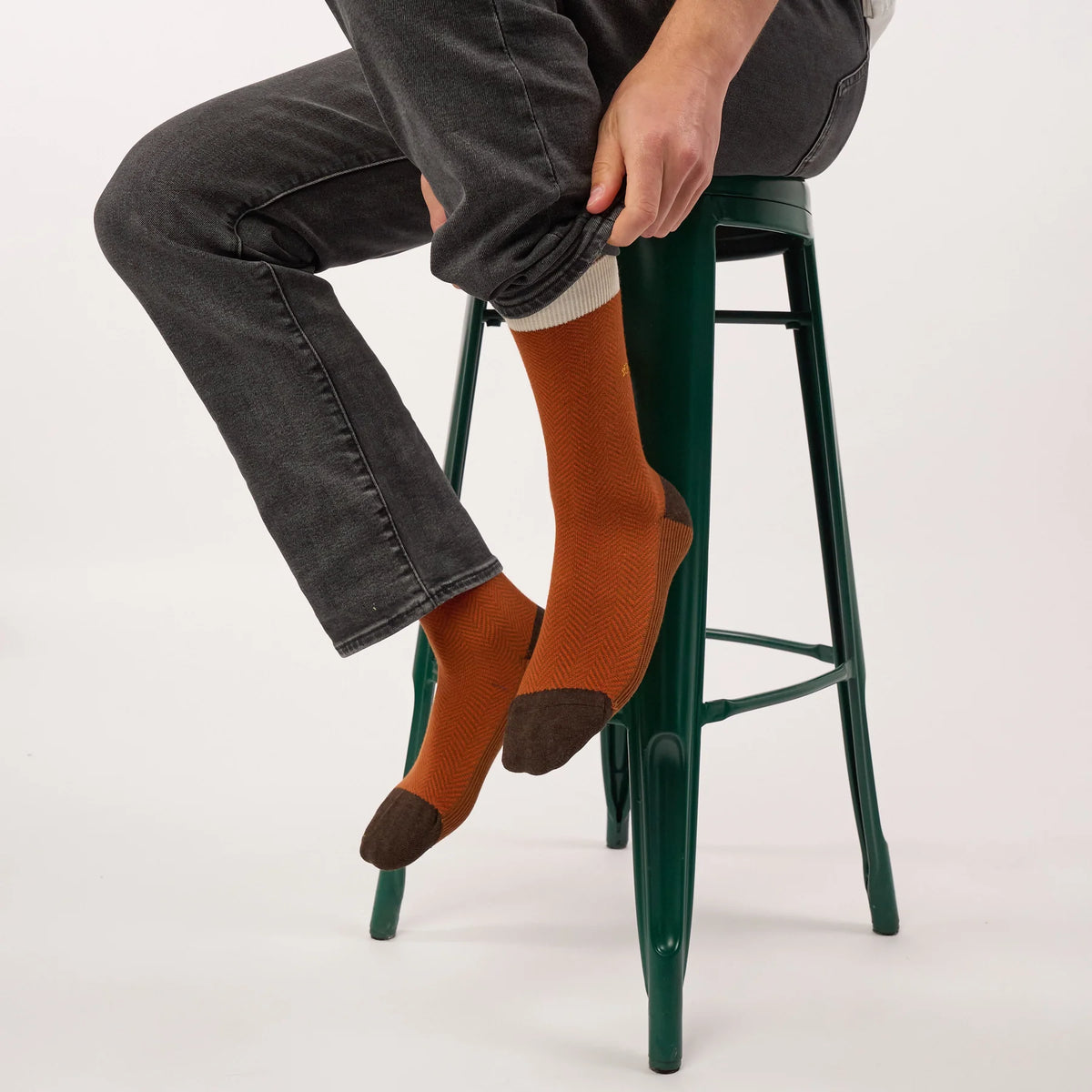 Chaussettes Chevrons Sépia portées avec un jean gris, assis sur un tabouret vert