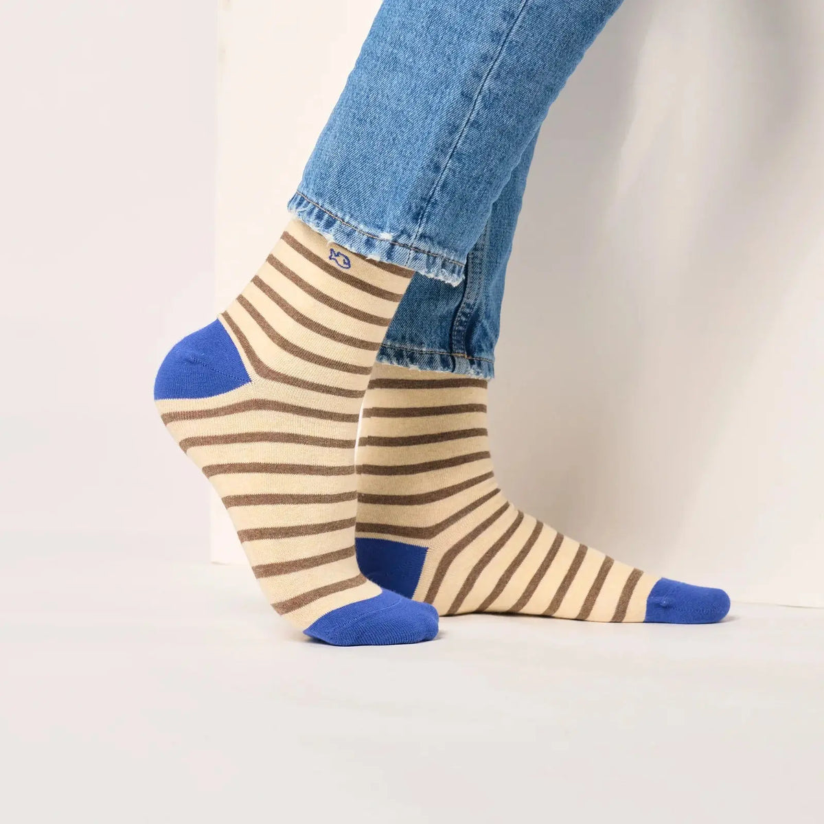 Chaussettes rayées beige et marron portées de côté, petit logo poisson brodé en bleu sous l'ourlet du jean.