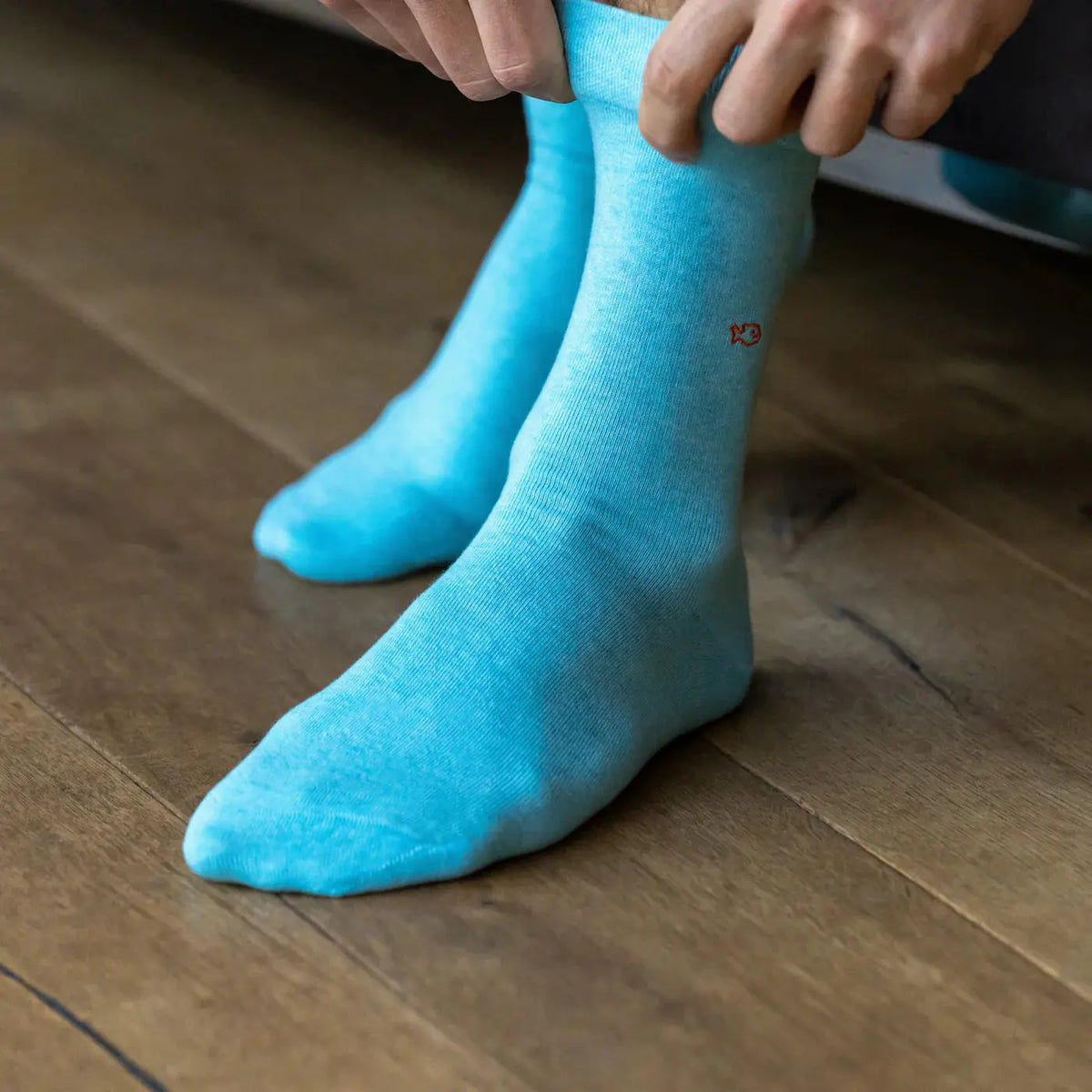 Gros plan sur la maille turquoise unie et son poisson brodé orange, bord-côte lisse en haut de tige.