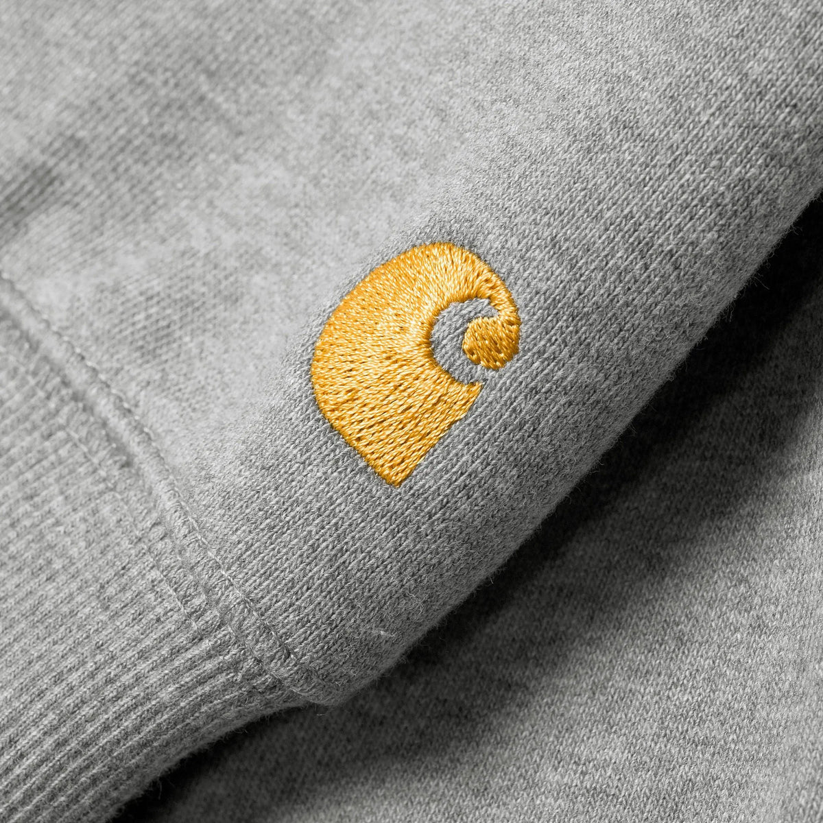 Macro du logo C brodé jaune doré sur la manche grise chinée du Hoodie Chase, au-dessus du poignet côtelé