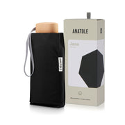 Mini parapluie Jane noir glissé dans sa housse, poignée en bois clair et dragonne rayée, coffret Anatole derrière