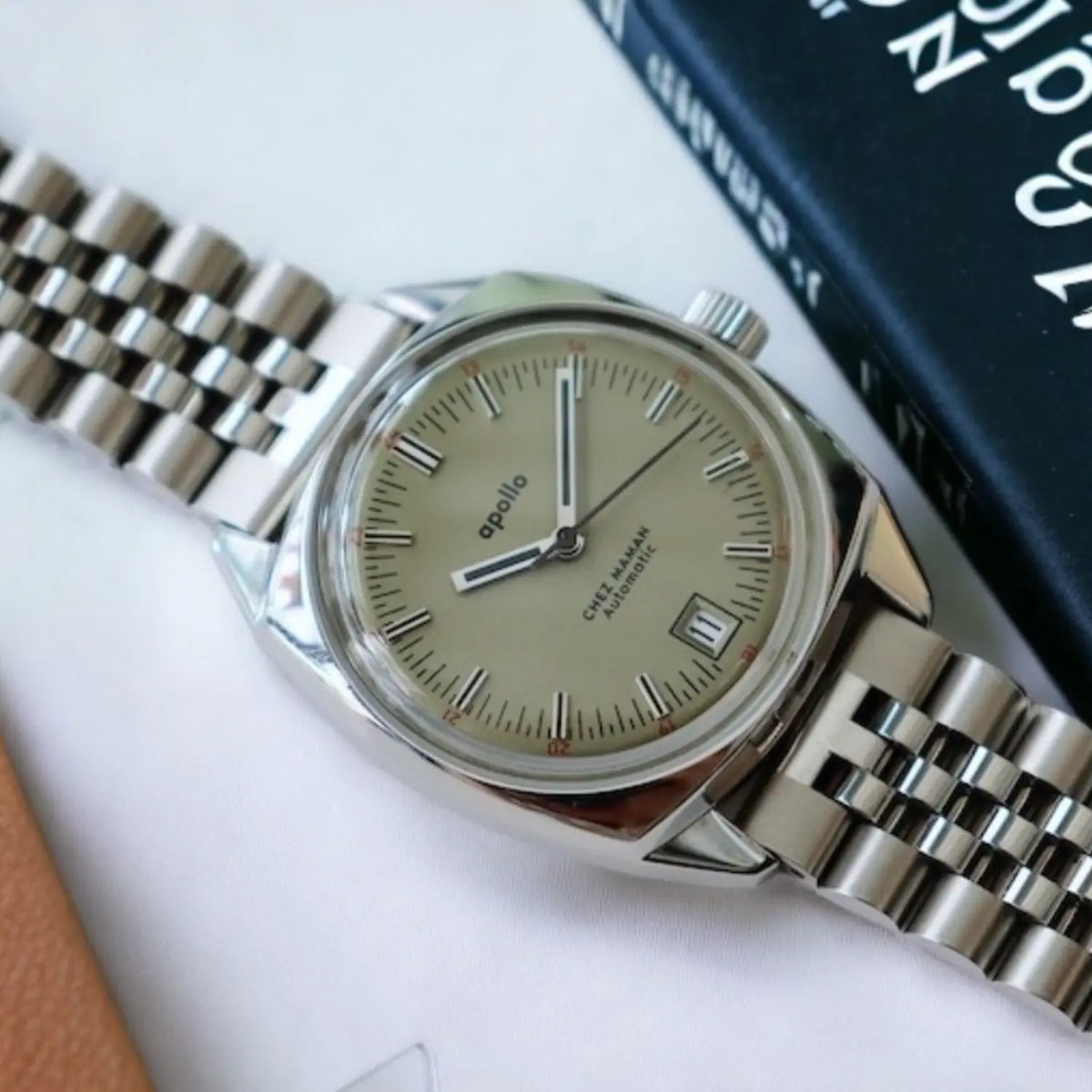 Montre Apollo posée près d'un livre, cadran sable clair, guichet de date à 4 h et bracelet jubilé acier