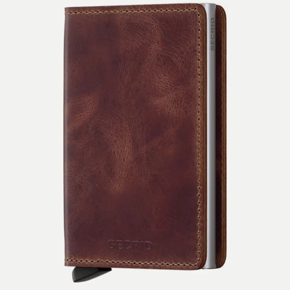 Portefeuille compact en cuir marron vieilli fermé, boîtier métal apparent sur la tranche et languette noire