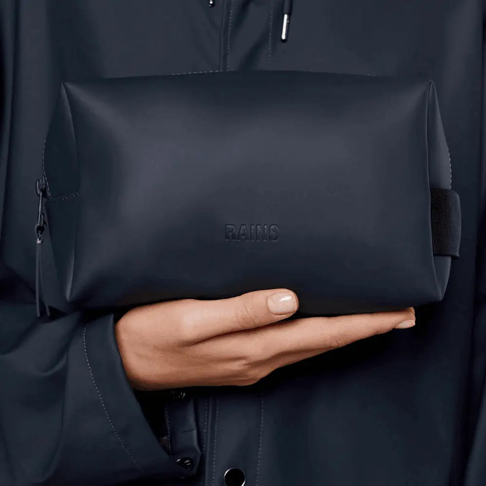 Wash Bag Small Rains bleu marine tenue en main devant une veste de pluie assortie, logo RAINS embossé bien visible